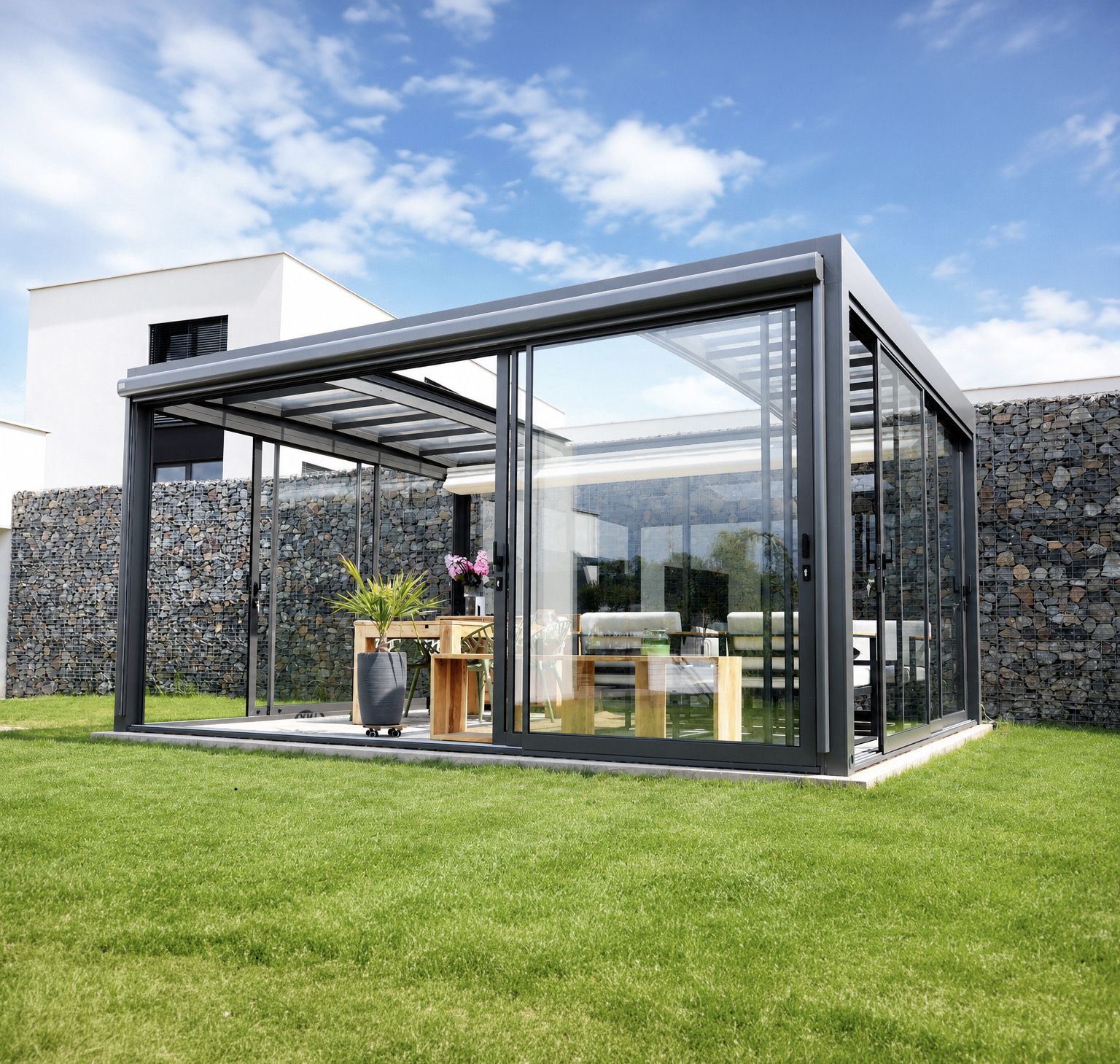 A glass garden room on a lawn with its door slid open, a gabion wall behind it